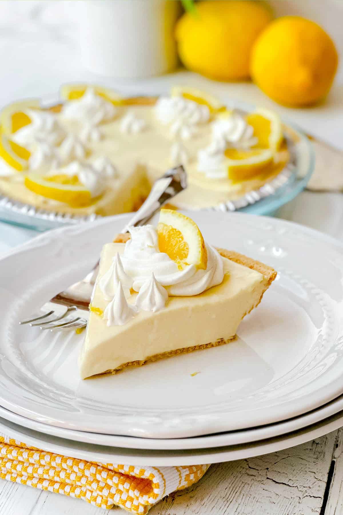 A slice of lemon pie with whipped topping and a lemon slice sits on a white plate; the whole lemon pie and fresh lemons are in the background.