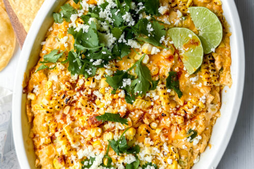 Top view of Mexican Street Corn Dip with fresh lime wedges, cilantro and Cotija cheese