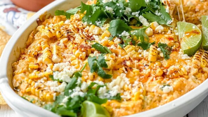A white baking dish filled with cheesy Mexican Street Corn Dip, topped with cilantro, cotija cheese, lime wedges, and served with tortilla chips on the side.