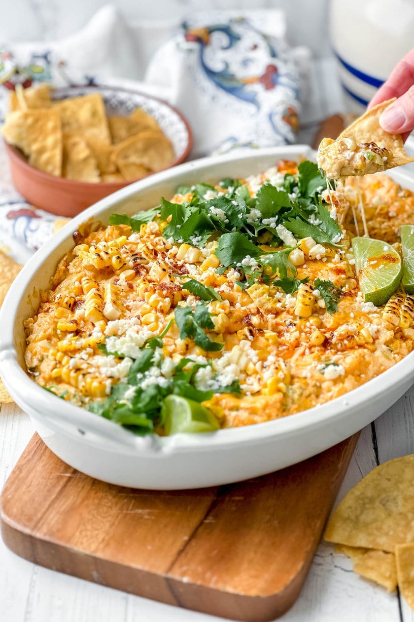 A white baking dish filled with cheesy Mexican Street Corn Dip, topped with cilantro, cotija cheese, lime wedges, and served with tortilla chips on the side.