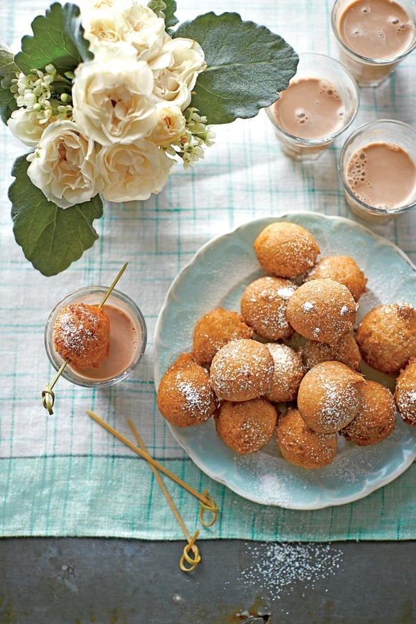 A plate of powdered sugar-dusted doughnut holes, made from quick and easy recipes, sits beside glasses of chocolate milk on a checkered tablecloth, with a bouquet of white flowers nearby.
