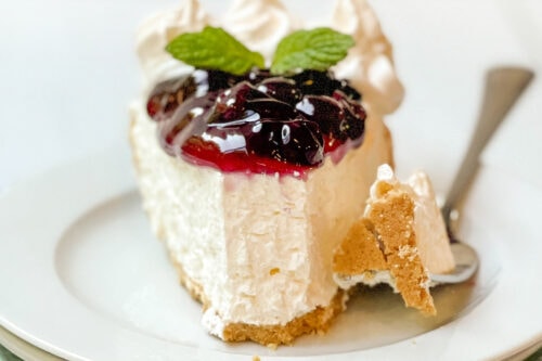 Front side view of 10 minute easy no-bake cheesecake topped with blueberry sauce, and fresh mint, with a bite missing on a white plate.