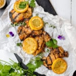 Grilled chicken thighs infused with a savory chicken marinade, topped with lemon slices and garnished with fresh herbs and edible flowers, arranged on crumpled parchment paper.