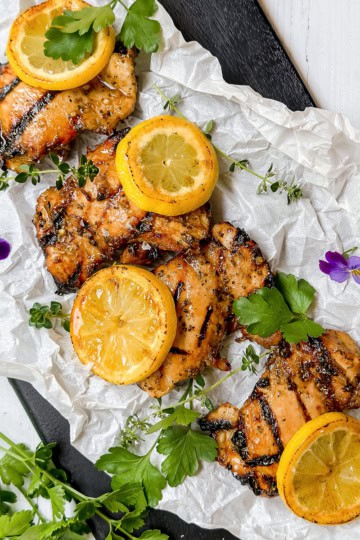 Grilled chicken thighs infused with a savory chicken marinade, topped with lemon slices and garnished with fresh herbs and edible flowers, arranged on crumpled parchment paper.