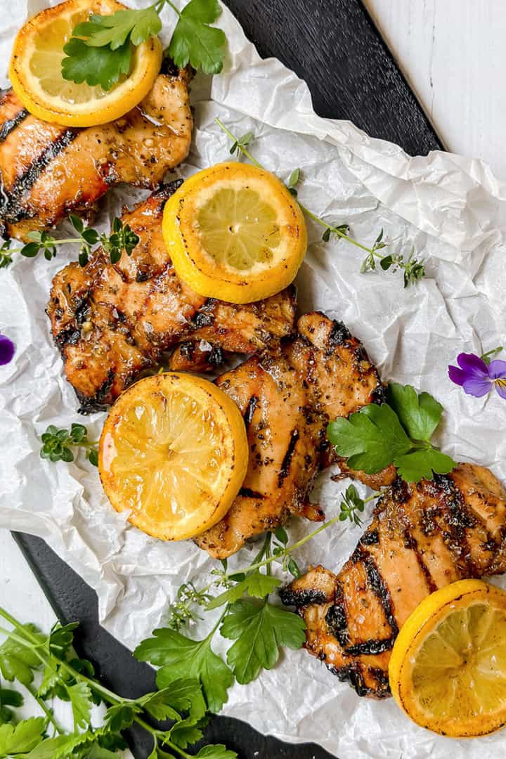 Grilled chicken thighs infused with a savory chicken marinade, topped with lemon slices and garnished with fresh herbs and edible flowers, arranged on crumpled parchment paper.