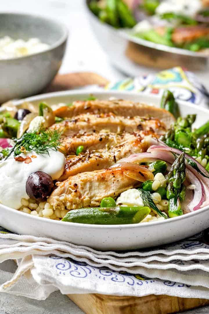 A bowl of Greek Chicken skillet slices on couscous with asparagus, snap peas, red onions, olives, a dollop of yogurt, and fresh herbs.