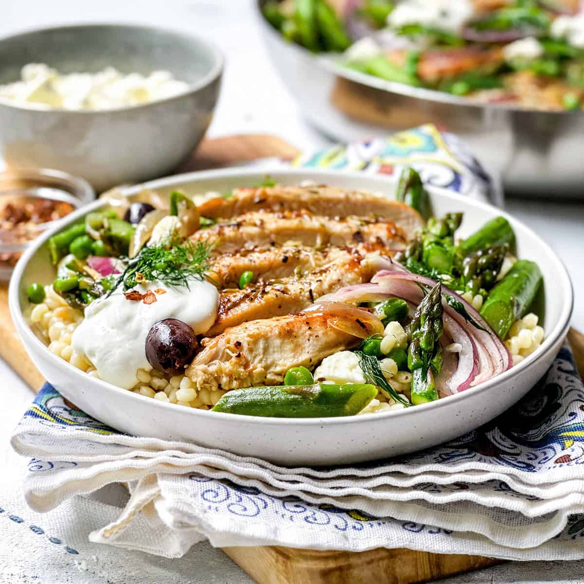 A bowl of Greek Chicken skillet slices on couscous with asparagus, snap peas, red onions, olives, a dollop of yogurt, and fresh herbs.