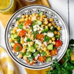 A colorful chickpea salad with cherry tomatoes, cucumber, green bell pepper, red onion, feta cheese, and fresh herbs is served on a decorative dish.
