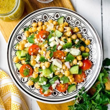 A colorful chickpea salad with cherry tomatoes, cucumber, green bell pepper, red onion, feta cheese, and fresh herbs is served on a decorative dish.