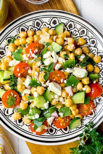 A colorful chickpea salad with cherry tomatoes, cucumber, green bell pepper, red onion, feta cheese, and fresh herbs is served on a decorative dish.