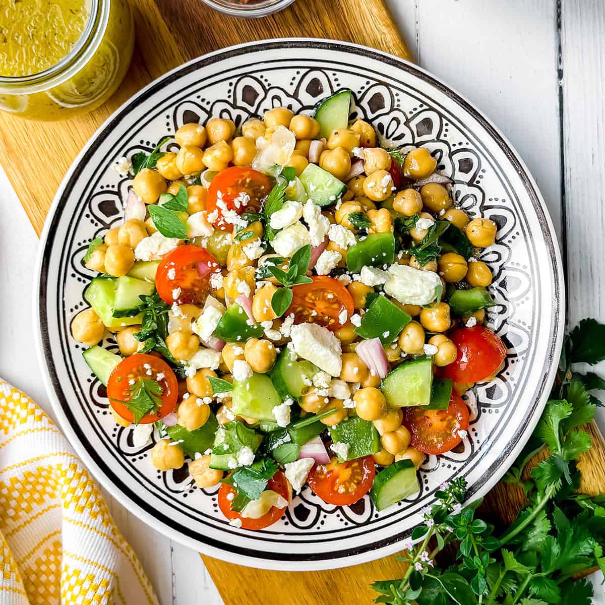 A colorful chickpea salad with cherry tomatoes, cucumber, green bell pepper, red onion, feta cheese, and fresh herbs is served on a decorative dish.