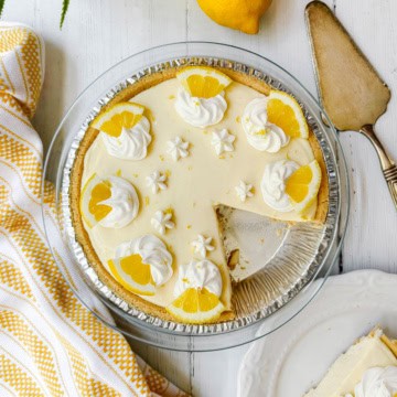 A lemon pie in a glass dish, topped with whipped cream and lemon slices, sits with one slice missing. A pie server and a napkin are nearby.