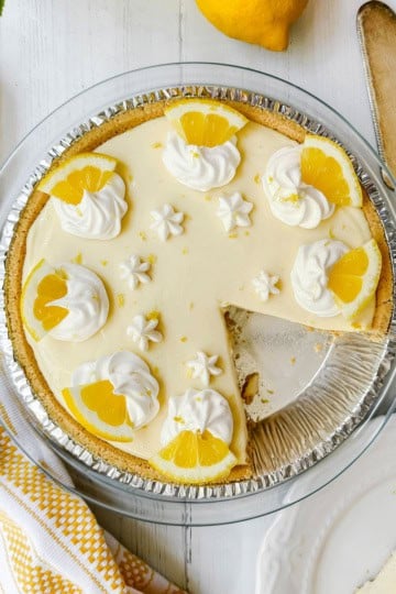 A lemon pie in a glass dish, topped with whipped cream and lemon slices, sits with one slice missing. A pie server and a napkin are nearby.