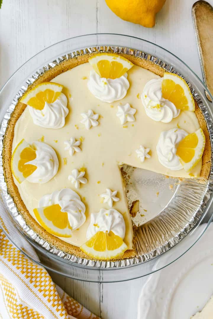A lemon pie in a glass dish, topped with whipped cream and lemon slices, sits with one slice missing. A pie server and a napkin are nearby.