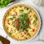 A baked vegetable quiche garnished with fresh microgreens sits in a white ceramic dish on a white wooden table, perfect for a delightful Mother's Day Breakfast, with crisp lettuce leaves visible nearby.