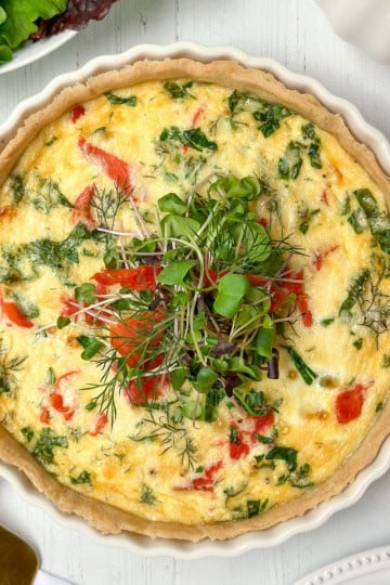 A baked vegetable quiche garnished with fresh microgreens sits in a white ceramic dish on a white wooden table, perfect for a delightful Mother's Day Breakfast, with crisp lettuce leaves visible nearby.