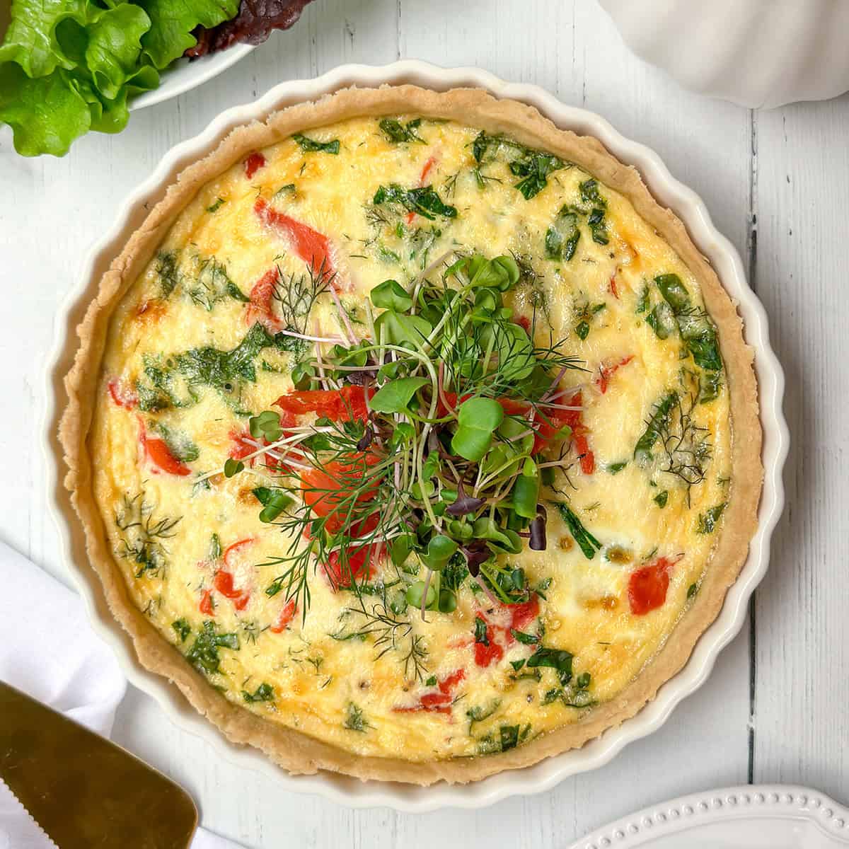 A baked vegetable quiche garnished with fresh microgreens sits in a white ceramic dish on a white wooden table, perfect for a delightful Mother's Day Breakfast, with crisp lettuce leaves visible nearby.