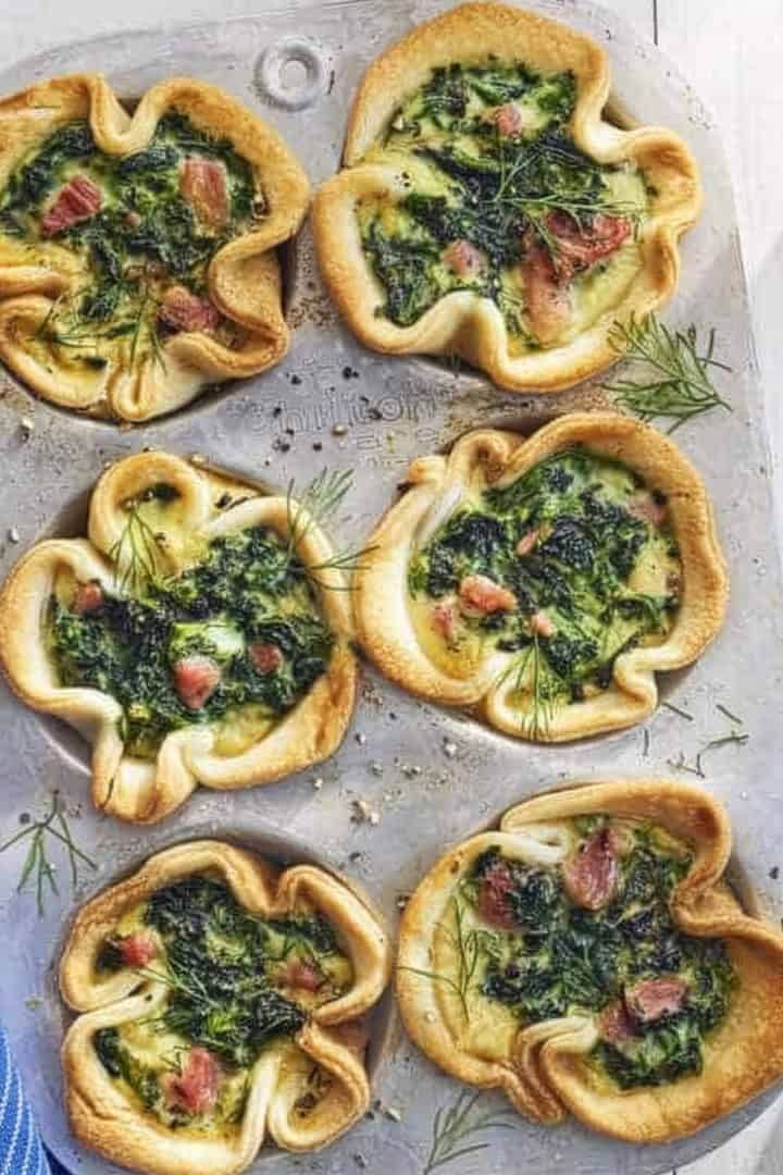 A muffin tin holds six baked mini quiches with leafy greens and bacon pieces, garnished with dill, perfect for a special Mother’s Day breakfast on a light-colored surface.