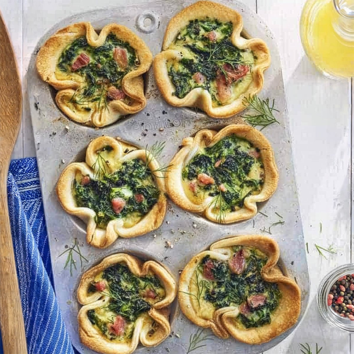 A muffin tin holds six baked mini quiches with leafy greens and bacon pieces, garnished with dill, perfect for a special Mother's Day breakfast on a light-colored surface.