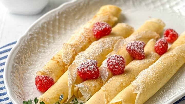 Four rolled Swedish pancakes topped with powdered sugar and raspberries are served on a white plate, with a cup of coffee and a small bowl of jam in the background.