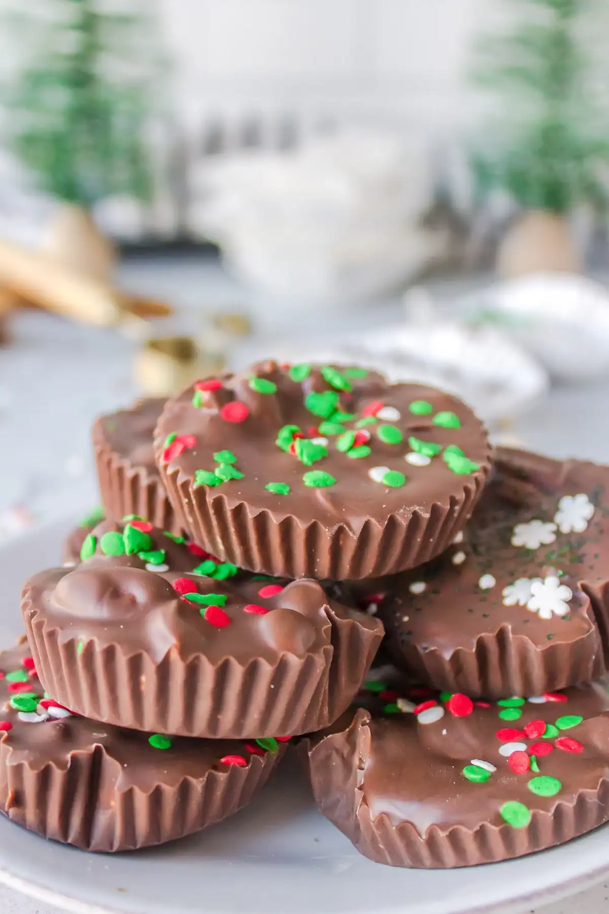 A stack of chocolate cups, resembling delightful Christmas candy, is decorated with colorful red, green, and white sprinkles. The treats are elegantly arranged on a white plate with blurred festive decorations in the background.