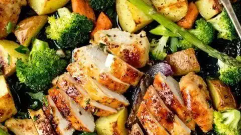 Sliced roasted chicken breast with assorted roasted vegetables—potatoes, carrots, broccoli, asparagus, and red onions—on a baking tray with a serving spoon. Perfect for May one pot dinners that make mealtime simple and delicious.