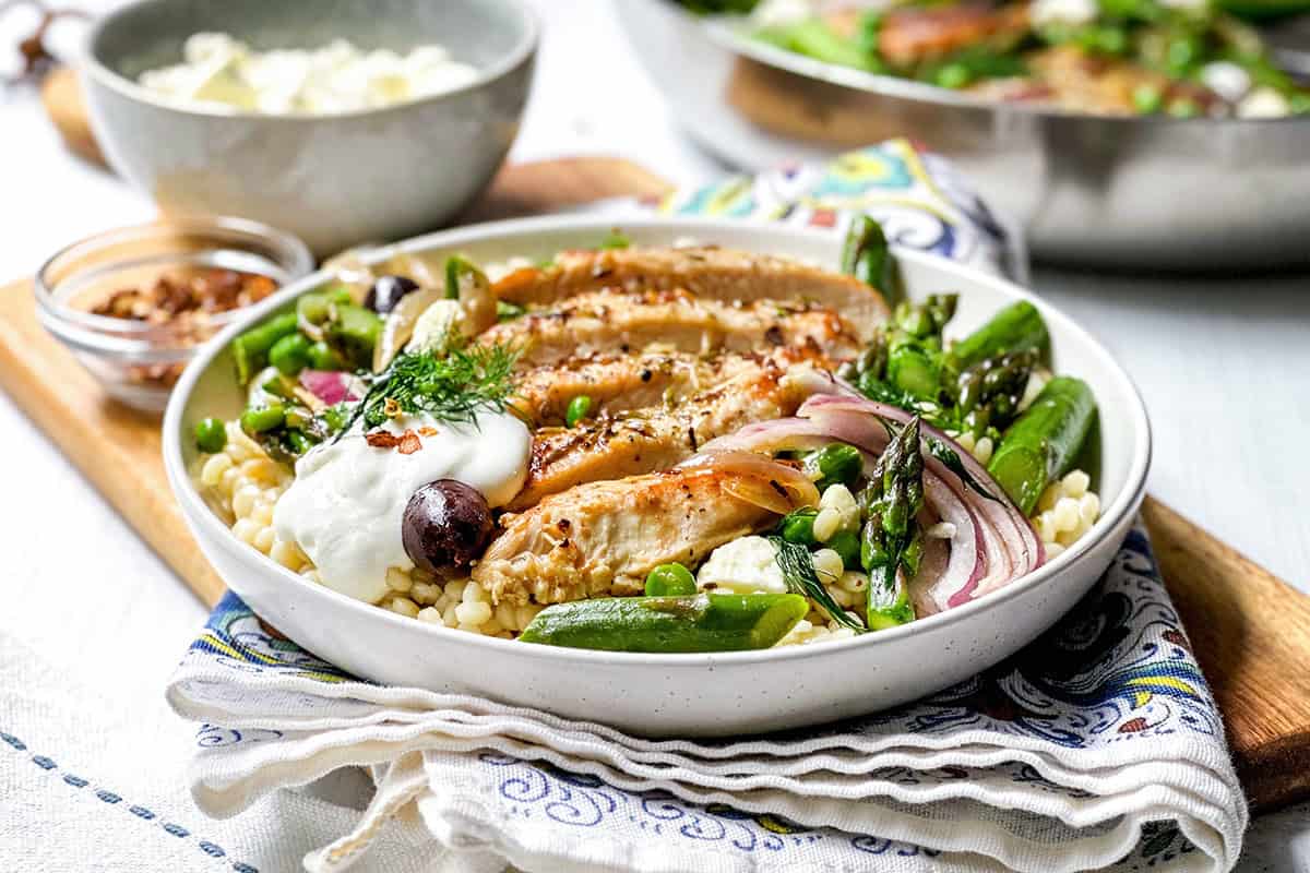 A bowl of couscous topped with Greek Chicken skillet, snap peas, asparagus, red onion, olives, yogurt, fresh herbs, and spices, with a bowl and skillet in the background.