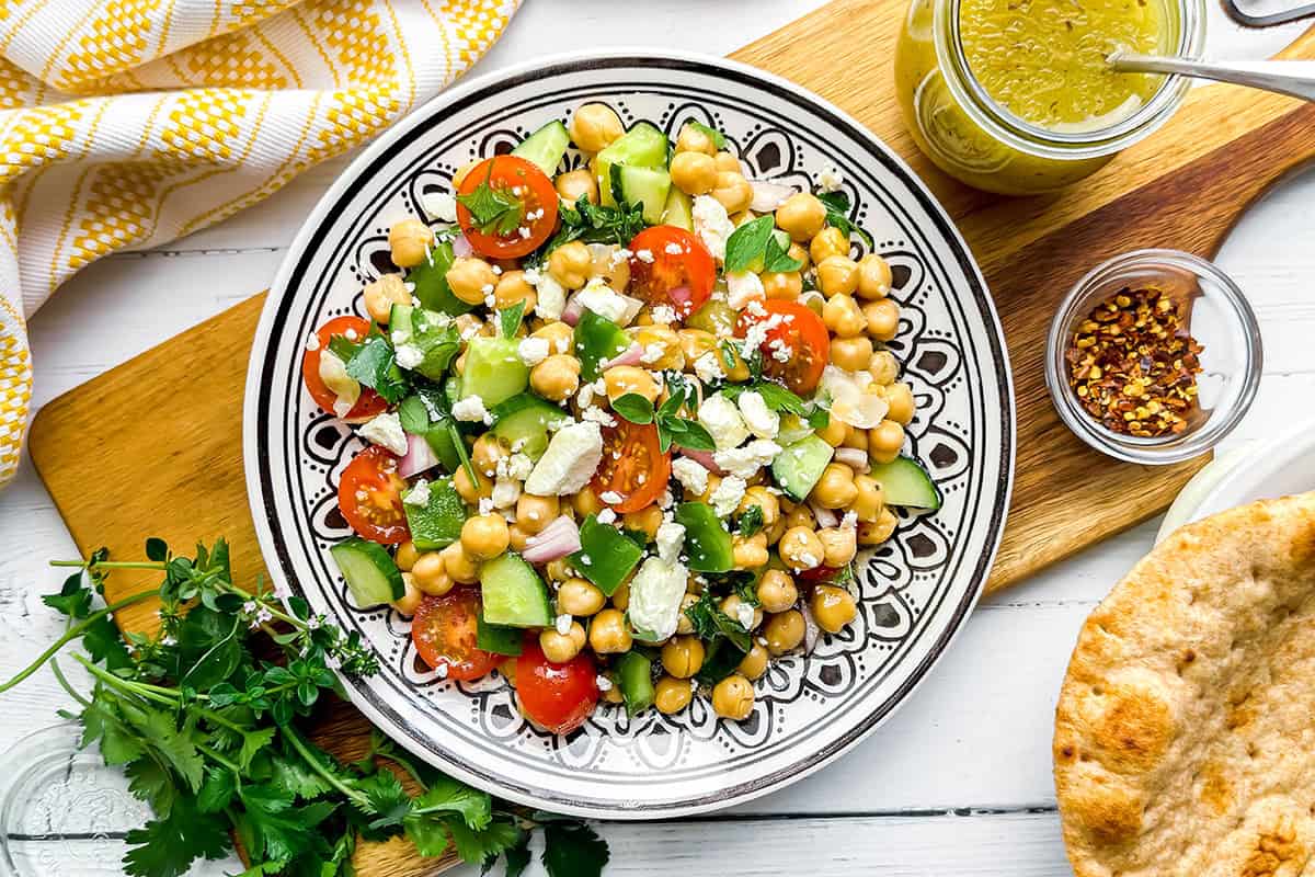 A colorful plate of chickpea salad with cherry tomatoes, cucumber, red onion, feta cheese, and herbs sits surrounded by pita bread, parsley, dressing, and red pepper flakes on a wooden table.