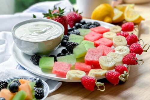 Side view of fruit skewers on a plate with yogurt dip and fresh strawberries