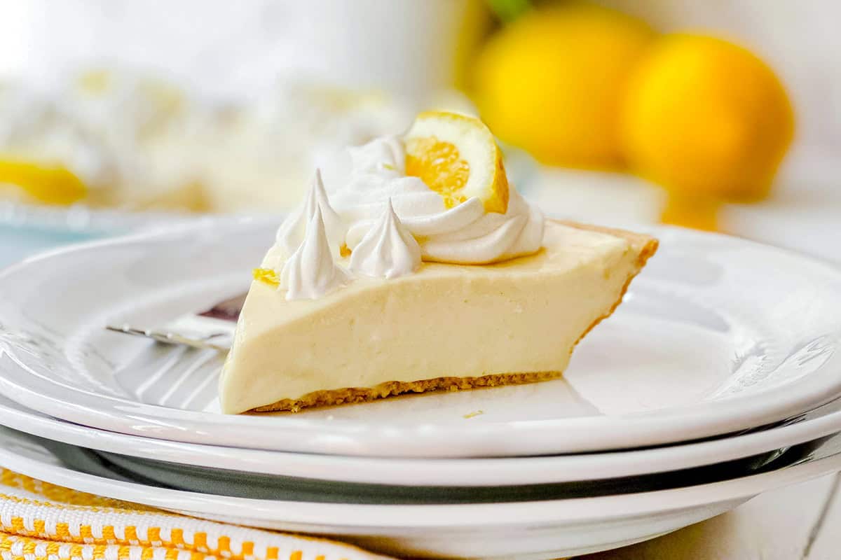 A slice of lemon pie with whipped topping and a lemon slice garnish sits on a white plate, while whole lemons add a fresh touch to the blurred background.