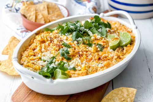 A white baking dish filled with Mexican Street Corn Dip, garnished with cilantro, cotija cheese, grilled corn, and lime wedges, surrounded by tortilla chips.