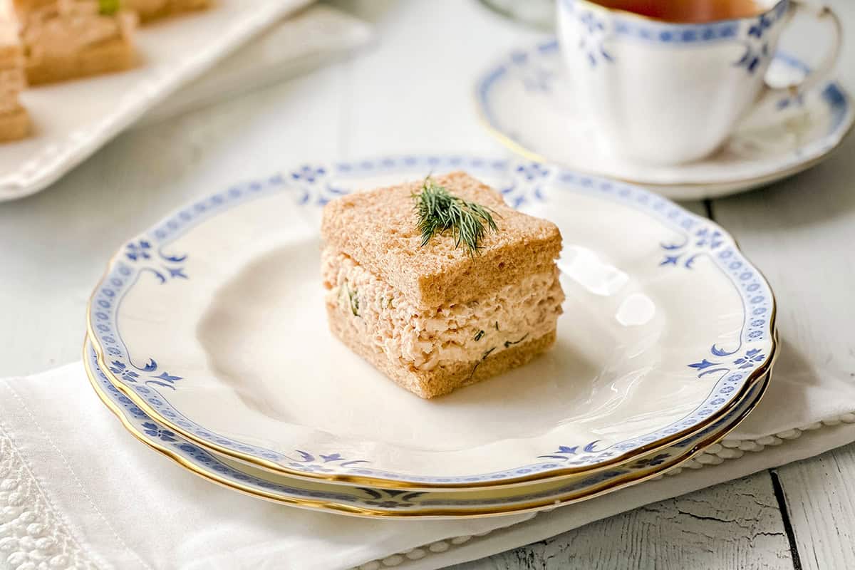 Salmon salad tea sandwich on whole wheat bread garnished with fresh dill and served with tea on blue-and-white china.