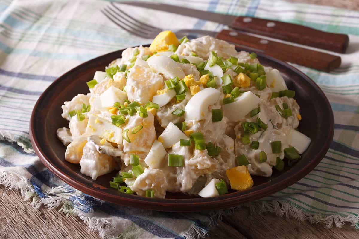 A plate of potato salad recipe with chopped boiled eggs and green onions, served on a dark plate with a fork and knife on a plaid cloth.