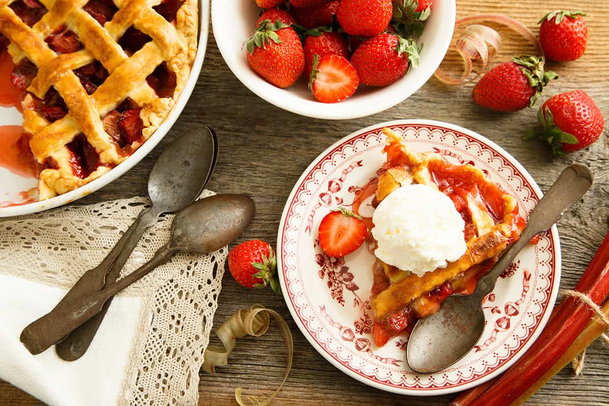 A slice of strawberry rhubarb pie with a scoop of vanilla ice cream on a plate, surrounded by whole strawberries, a bowl of strawberries, and vintage spoons on a wooden table.