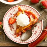 A slice of strawberry rhubarb pie topped with vanilla ice cream sits on a decorative plate, surrounded by a spoon, fresh strawberries, and rhubarb.