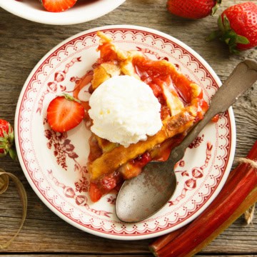 A slice of strawberry rhubarb pie topped with vanilla ice cream sits on a decorative plate, surrounded by a spoon, fresh strawberries, and rhubarb.