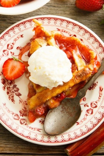 A slice of strawberry rhubarb pie topped with vanilla ice cream sits on a decorative plate, surrounded by a spoon, fresh strawberries, and rhubarb.