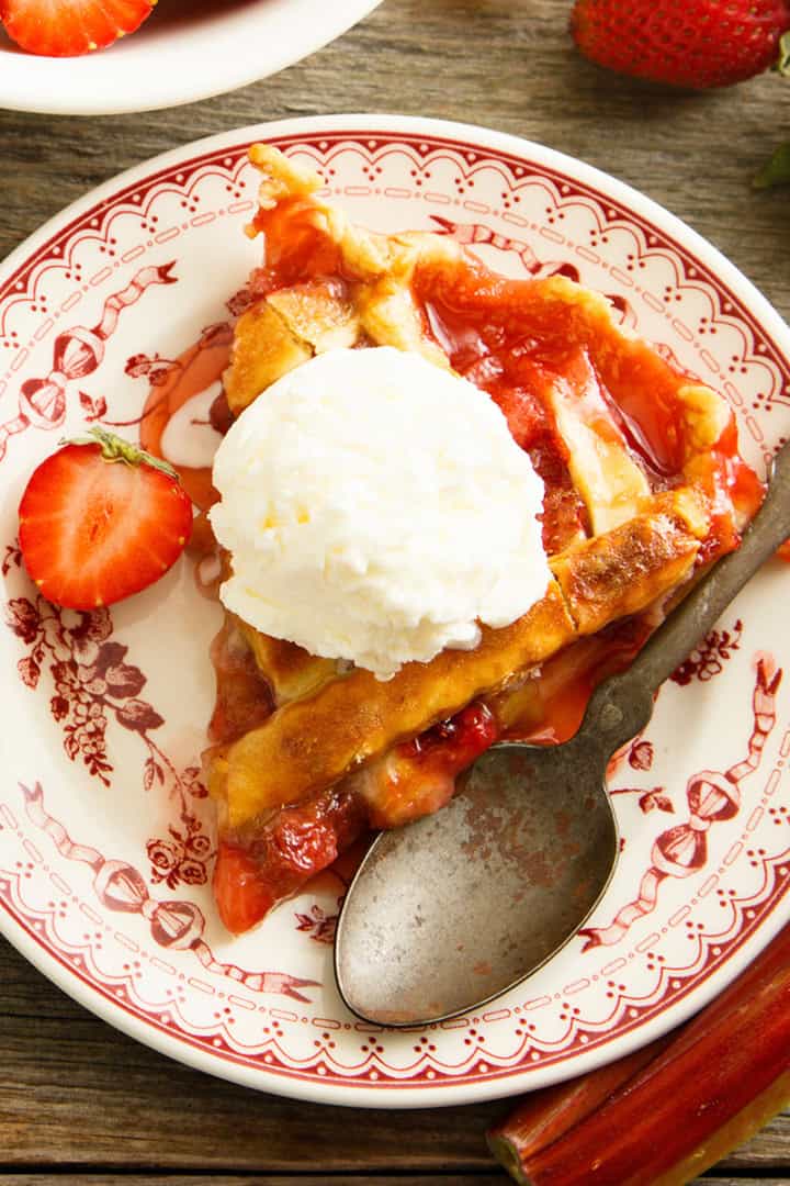 A slice of strawberry rhubarb pie topped with vanilla ice cream sits on a decorative plate, surrounded by a spoon, fresh strawberries, and rhubarb.