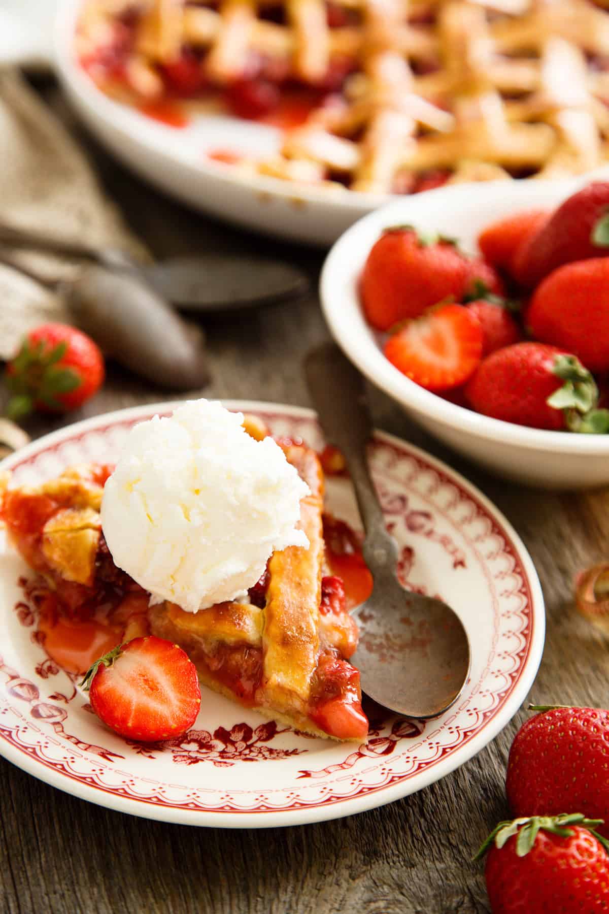 A slice of strawberry rhubarb pie topped with a scoop of vanilla ice cream sits on a plate beside a spoon, with fresh strawberries and more pie in the background.