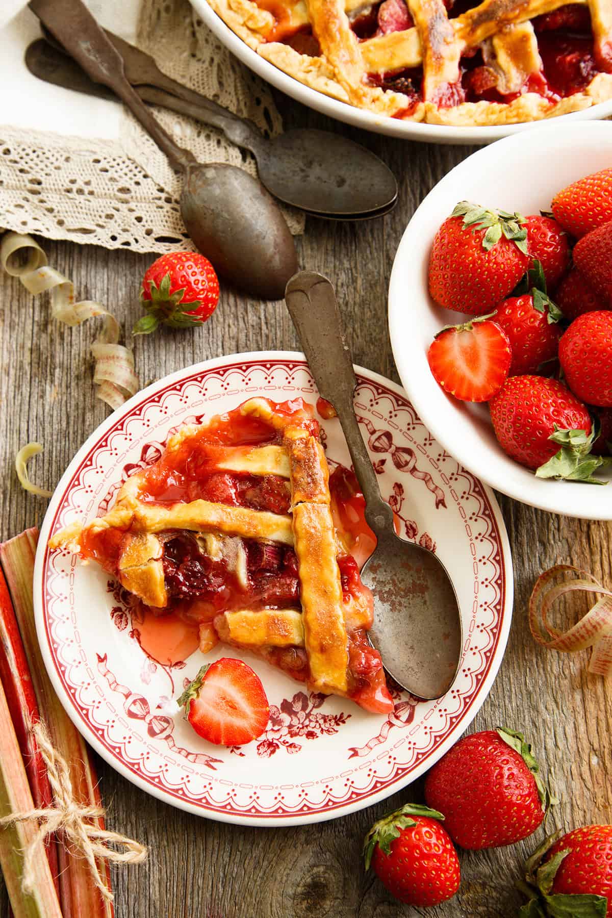 A slice of strawberry rhubarb pie sits on a decorative plate with a spoon, surrounded by fresh strawberries and a bowl of strawberries on a rustic wooden table.