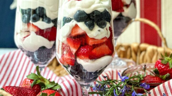Three glass parfaits layered with whipped cream, blueberries, and strawberries sit in a woven tray decorated with fresh strawberries, flowers, and a striped cloth. A flag background adds a festive touch to the parfait presentation.