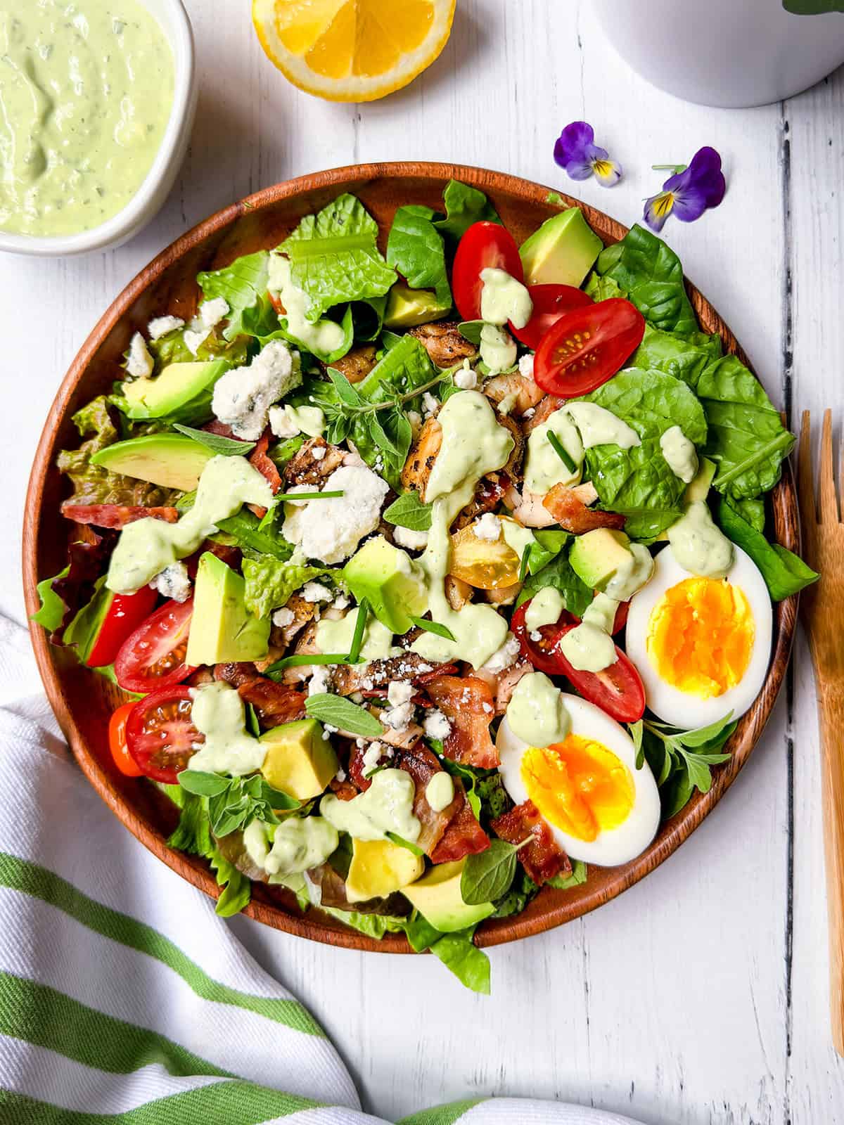 A wooden bowl of cobb salad with chicken, greens, tomatoes, avocado, blue cheese, crumbled bacon, and a halved soft-boiled egg, topped with creamy avocado dressing.
