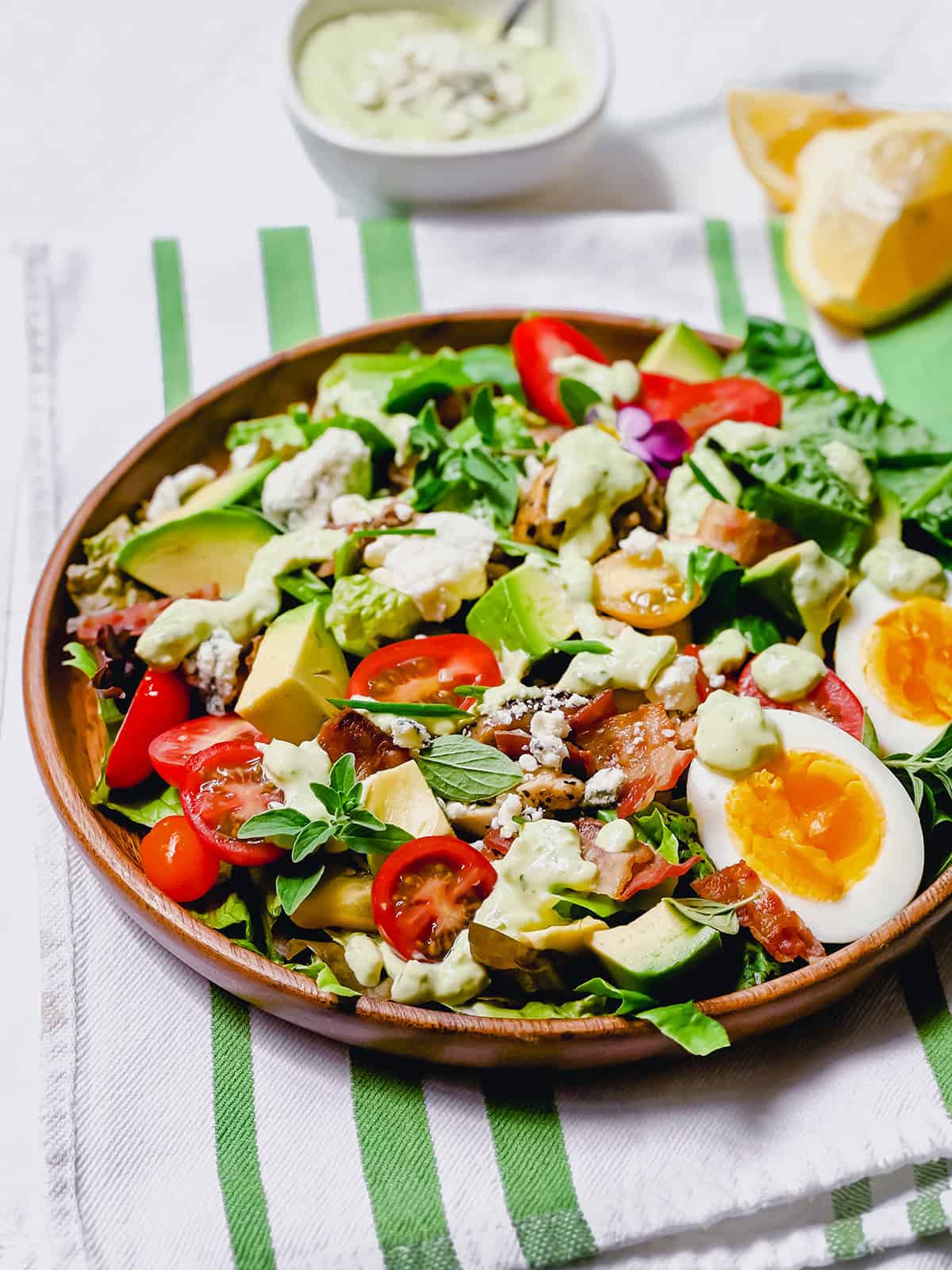 A wooden plate with a cobb salad featuring avocado, cherry tomatoes, greens, blue cheese, bacon, chicken, and a soft-boiled egg, served on a striped napkin with dressing and lemon on the side.