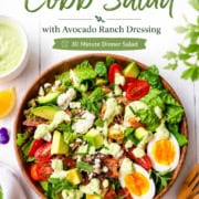 A bowl of chicken Cobb salad with eggs, avocado, tomatoes, lettuce, bacon, and avocado ranch dressing sits surrounded by a napkin, fork, flowers, and an extra bowl of dressing—a perfect classic Cobb salad setup.