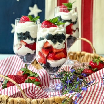 Three parfait glasses filled with layers of yogurt, strawberries, and blueberries are arranged in a basket with fresh strawberries and flowers, set in front of an American flag for a festive parfait display.