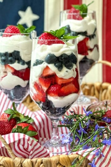 Three parfait glasses filled with layers of yogurt, strawberries, and blueberries are arranged in a basket with fresh strawberries and flowers, set in front of an American flag for a festive parfait display.