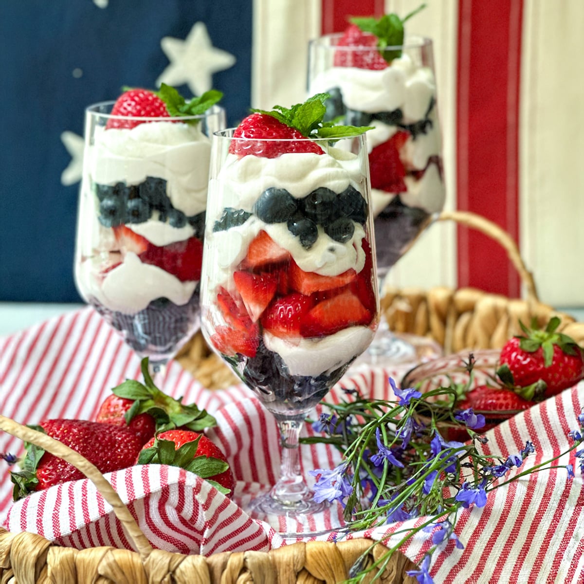 Three parfait glasses filled with layers of yogurt, strawberries, and blueberries are arranged in a basket with fresh strawberries and flowers, set in front of an American flag for a festive parfait display.