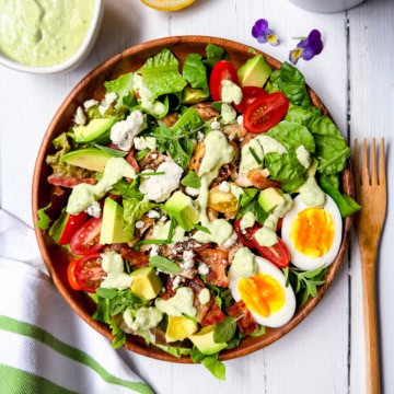 A wooden bowl filled with cobb salad-leafy greens, cherry tomatoes, avocado, bacon, feta cheese, a sliced boiled egg, and creamy dressing-sits on a white table with a fork and napkin beside it.