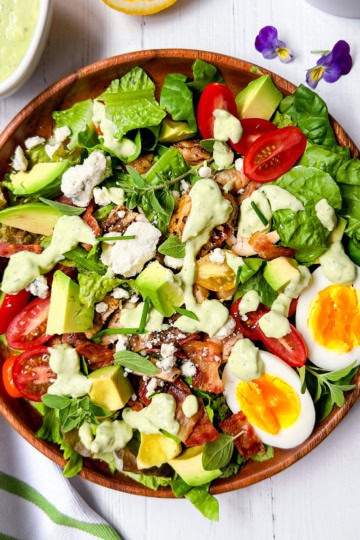 A wooden bowl filled with cobb salad—leafy greens, cherry tomatoes, avocado, bacon, feta cheese, a sliced boiled egg, and creamy dressing—sits on a white table with a fork and napkin beside it.