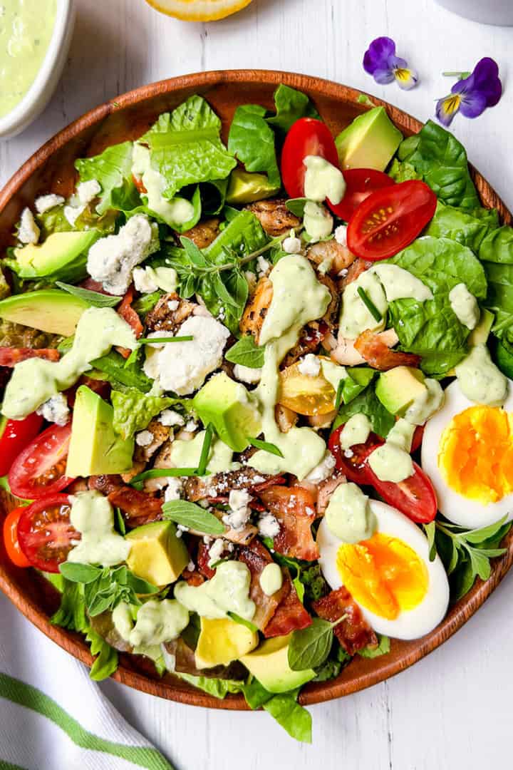 A wooden bowl filled with cobb salad—leafy greens, cherry tomatoes, avocado, bacon, feta cheese, a sliced boiled egg, and creamy dressing—sits on a white table with a fork and napkin beside it.